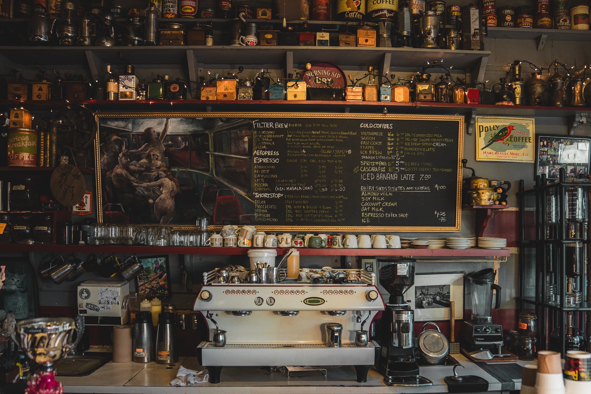Head photo of cappuccino bar at our coffee shop The Roasterie. The surroundings look like a coffee museum, you can see coffee grinders on shelves, old coffee makers, porcelain cups, beautiful white espresso machine making the creamiest espresso shots.