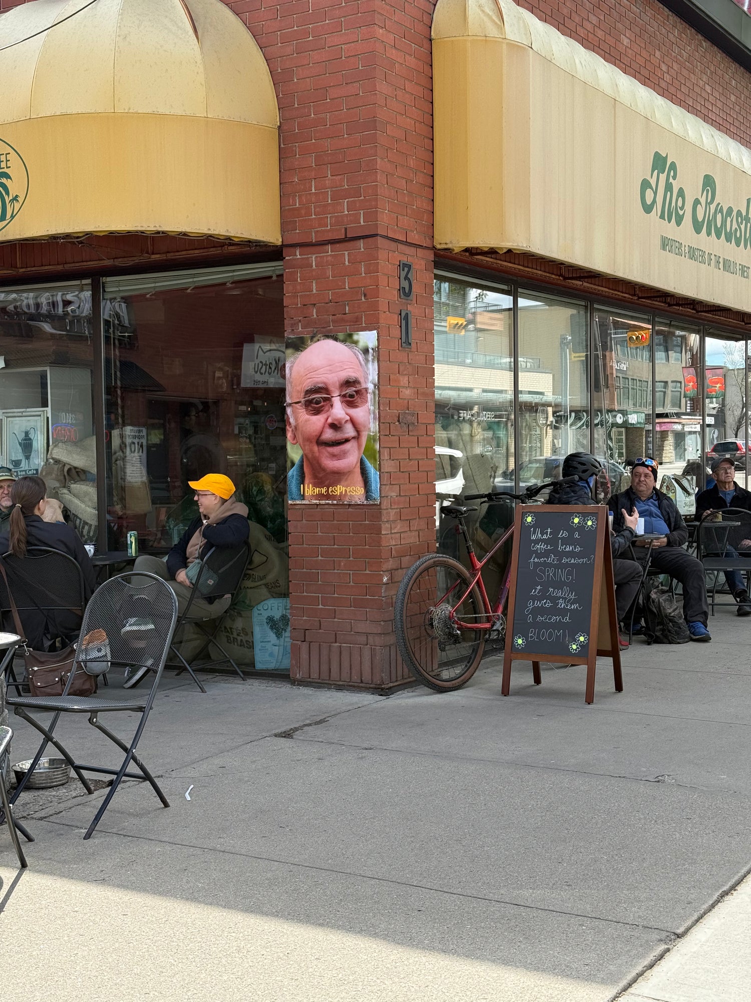Outdoor seating area in front of a brick building with people sitting and a poster on the wall. local-calgary-cafe-kensington-patio